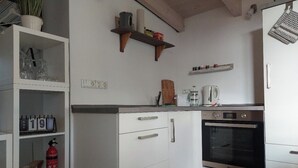 A modern kitchen with white cabinetry, a built-in oven, and a shelf with kitchen utensils. - Small, fine & rural (Pronstorf)