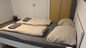Una cama individual con colchón gris y almohadas color beige, una mesita de noche con lámpara, y una puerta de madera al fondo.