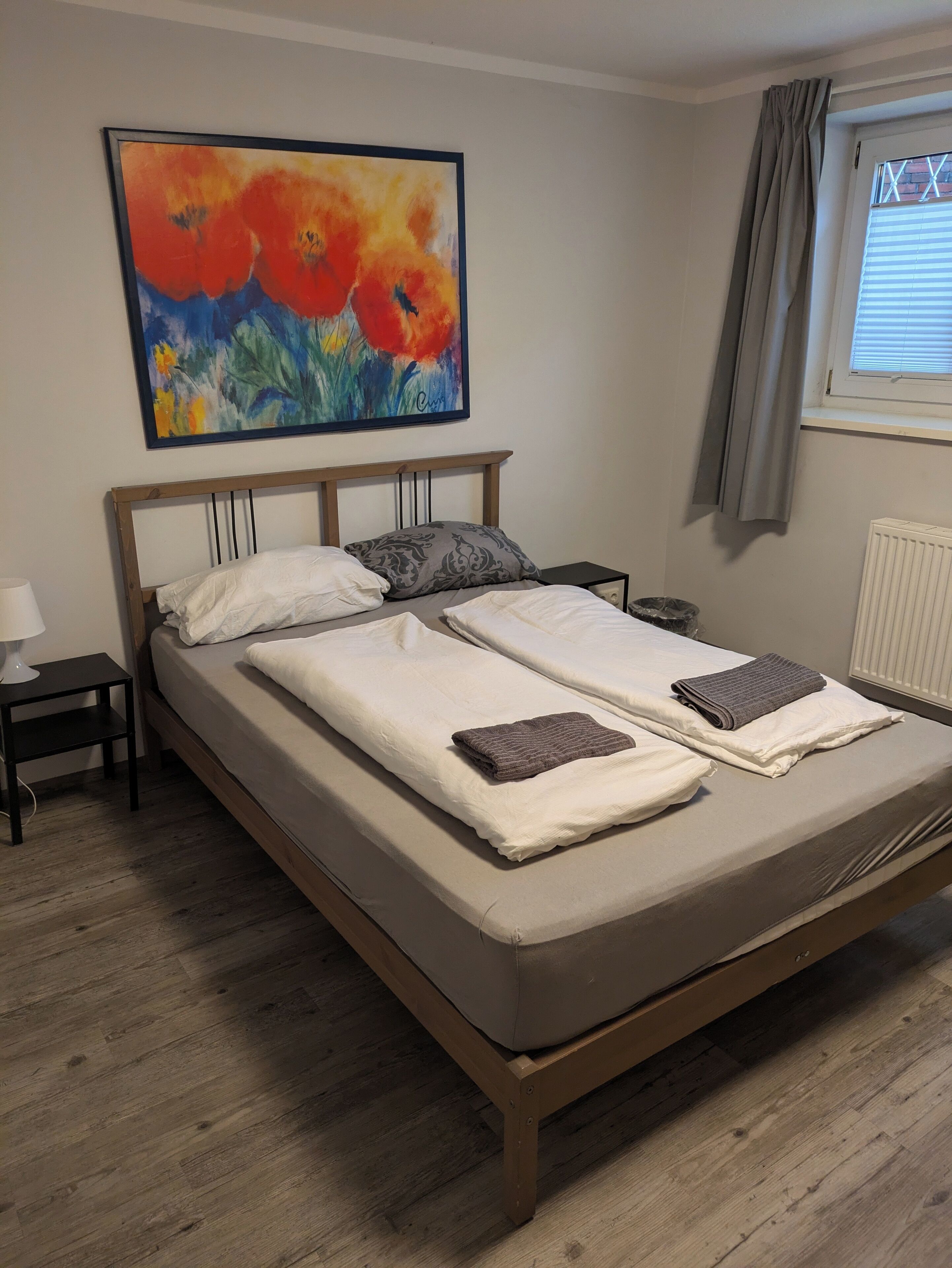 Un lit double avec deux oreillers et une couverture, une table de chevet avec une lampe, un tableau au mur et une fenêtre avec des rideaux.