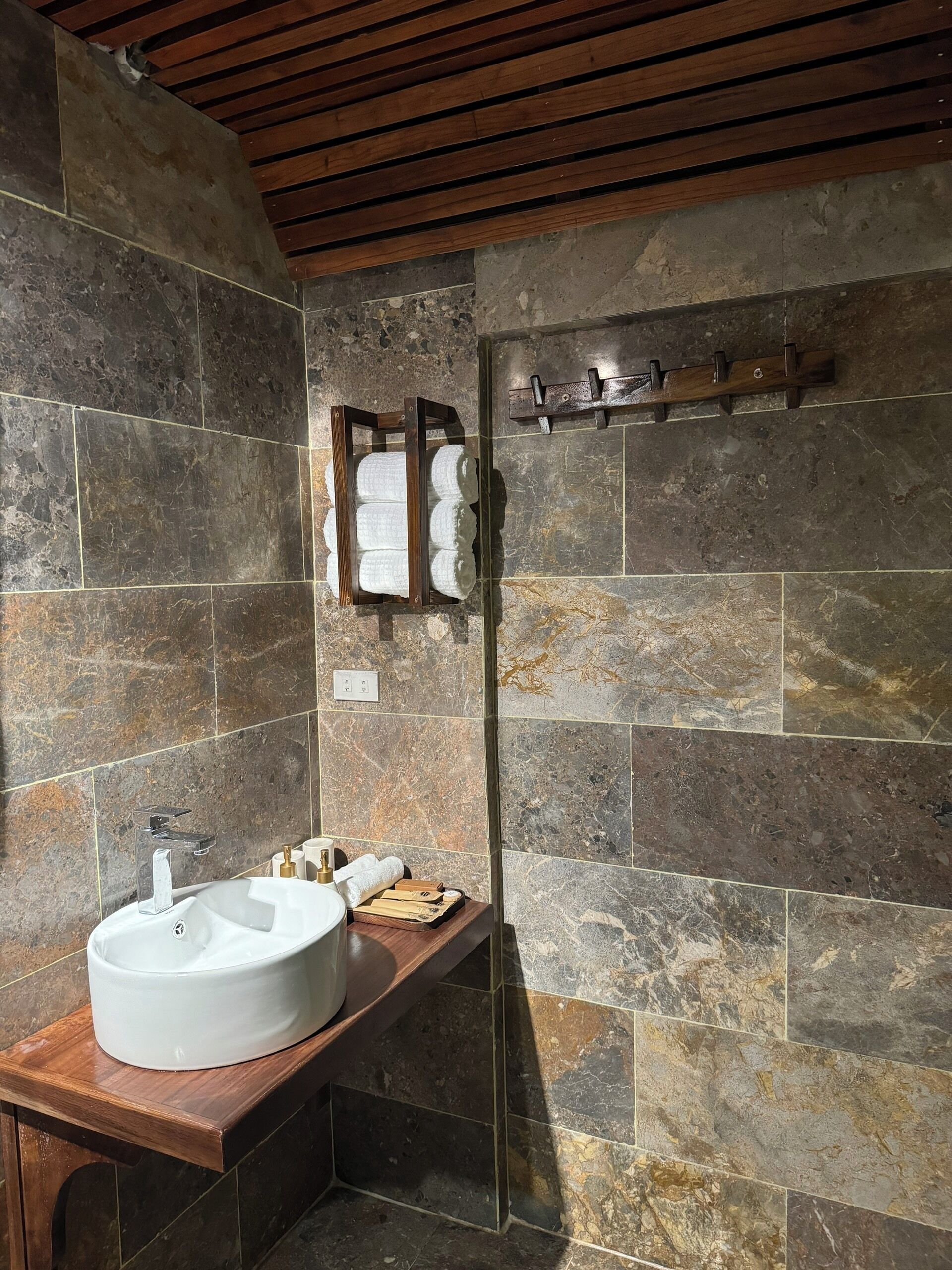 A bathroom with a stone wall, a wooden bench, a sink, and a glass shower enclosure.