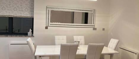 Un coin repas avec une table et des chaises blanches, un grand miroir et un luminaire suspendu.