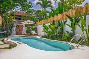 A swimming pool with a wooden fence, lounge chairs, and a small building with a tiled roof. - Malta Villa by Betterplace (Ubud)