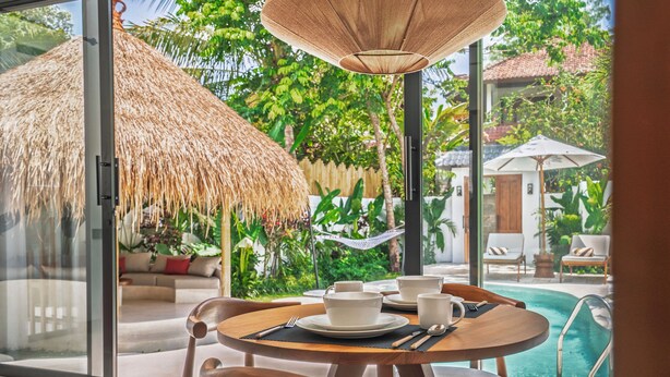 A dining area with a table set for two, a thatched umbrella, and a view of a swimming pool and lush greenery. - Malta Villa by Betterplace (Ubud)