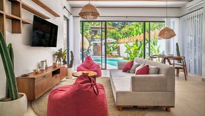 A modern living room with a sofa, red bean bag chairs, a TV mounted on the wall, and a large window overlooking a garden and pool. - Malta Villa by Betterplace (Ubud)