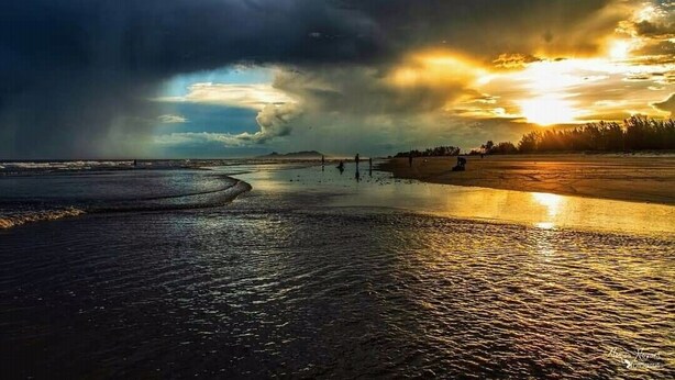 Een strand met een ondergaande zon, kalm water en een verre kustlijn.
