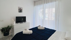 Ein modernes Schlafzimmer mit einem Bett, einem Fernseher auf einer weißen Kommode, einer Topfpflanze und einem Fenster mit transparenten Vorhängen.