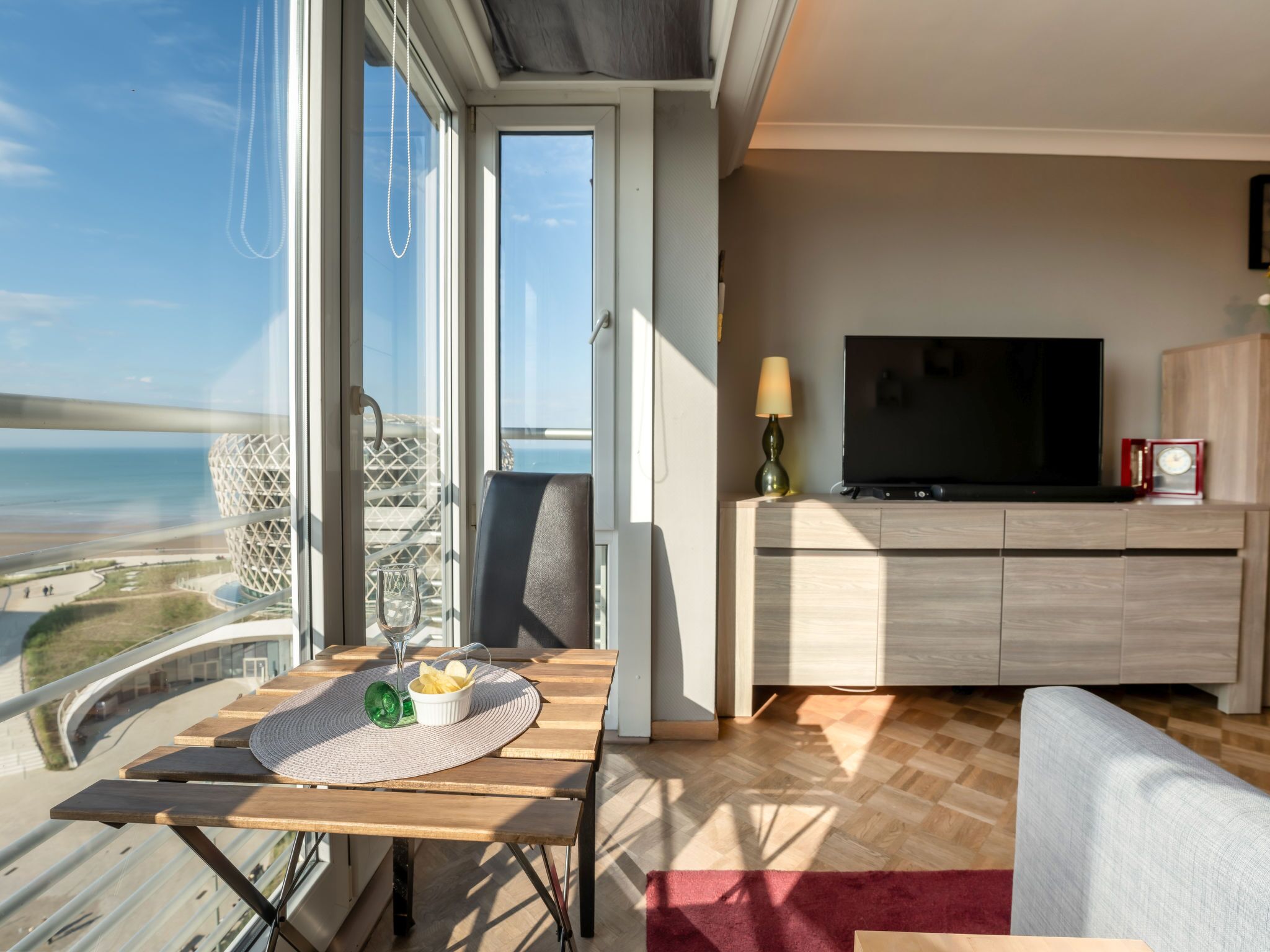 Ein modernes Wohnzimmer mit großem Fernseher, einem Esstisch mit Tischdecke und Glas sowie Blick auf den Strand und eine ungewöhnliche Bauweise.