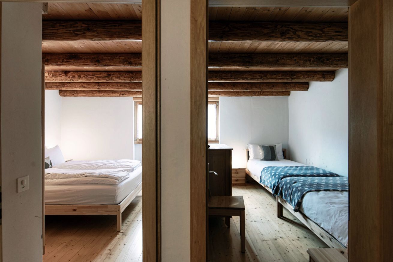 Een kamer met een houten vloer, een bed met een witte matras, een houten bankje en een bureau met een stoel.