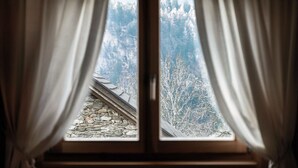 Una ventana con cortinas que muestra un tejado de piedra y una vista de la montaña boscosa.