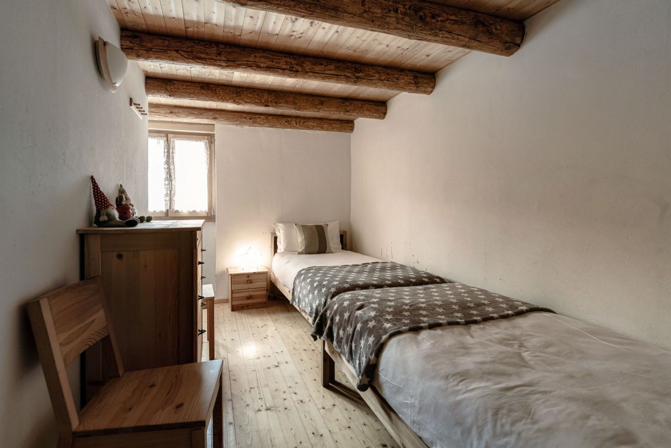 Um quarto pequeno e acolhedor com teto de madeira, uma cama de solteiro, uma mesa de cabeceira, uma cadeira de madeira e uma pequena mesa.