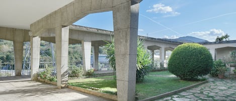 A covered outdoor area with stone columns, a paved walkway, and a small garden with a neatly trimmed bush.