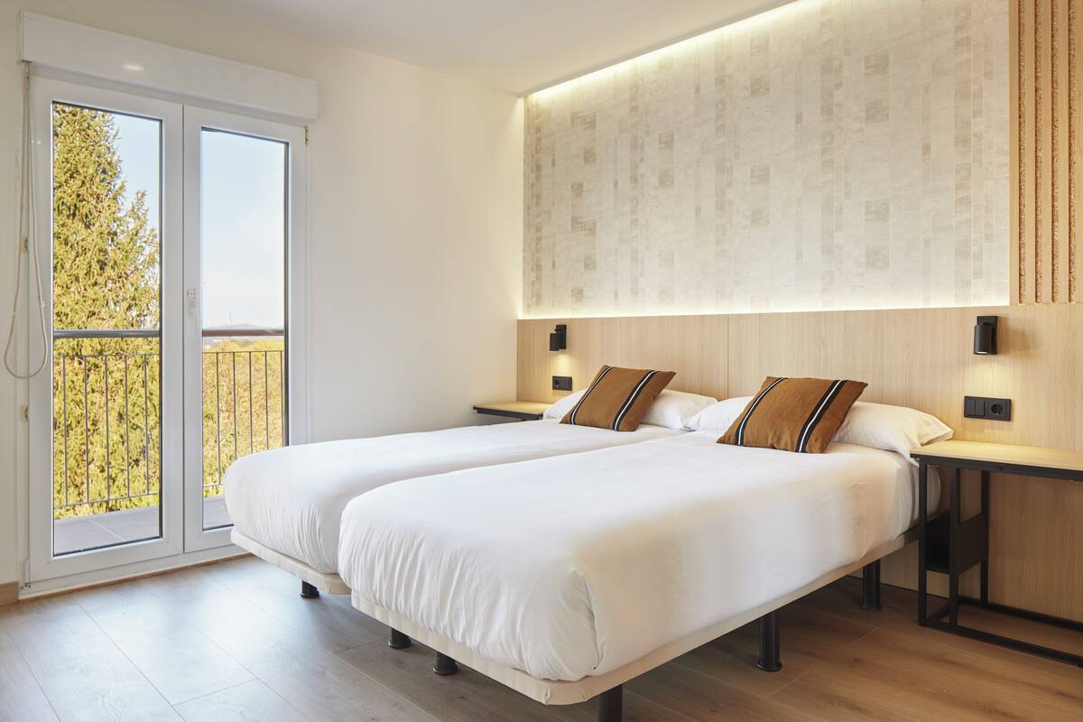 A modern hotel room with two beds, a large window with a view of trees, and a wall-mounted light fixture.