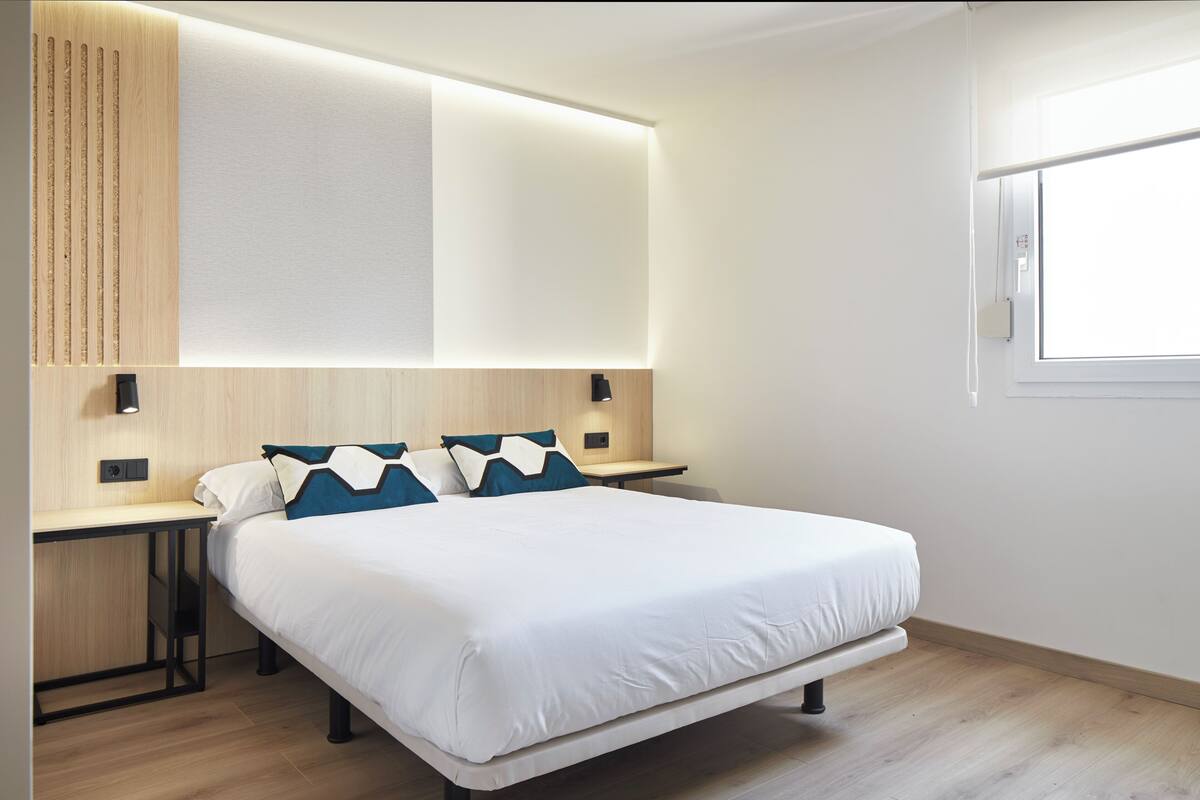 A modern bedroom with a large bed, a desk, and a wall-mounted light fixture.