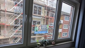 Ein Fenster mit Blick auf ein Baugerüst, ein Schild, das eine Stelle bewirbt, und eine Heizung darunter.