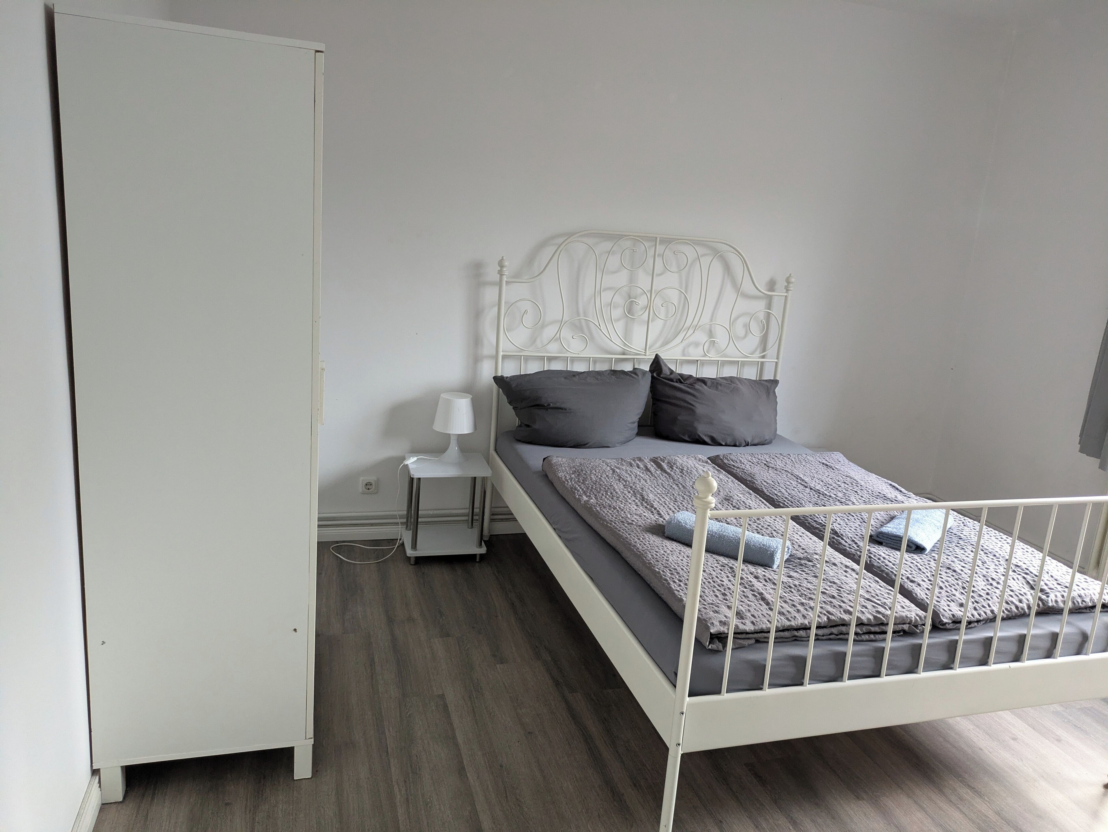 Quarto com estrutura de cama de metal branco, mesa de cabeceira com abajur e guarda-roupa.