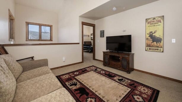 A living room with a large TV, a sofa, a patterned rug, and a poster of a moose. - Hot Tub | Mountain Views | Steps to Trails (Winter Park)