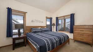 A bedroom with a large bed, two windows with curtains, a wooden dresser, and a nightstand with a lamp.