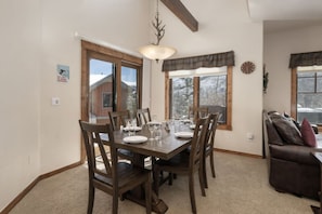 Comedor con mesa de madera dispuesta para cuatro personas, sillas, una araña y ventanas con cortinas.