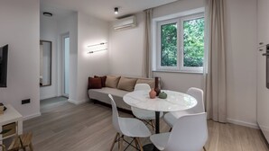 Una sala de estar moderna con una mesa de comedor redonda, sillas blancas, un sofá color beige, un televisor de pantalla plana y un amplio ventanal con cortinas.