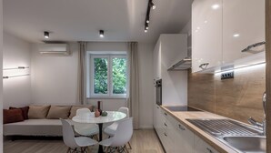 Una cocina moderna con encimera de madera, pileta de acero inoxidable y horno empotrado. Hay un comedor con una mesa redonda y sillas blancas. Un ventanal ofrece luz natural y vistas al exterior.