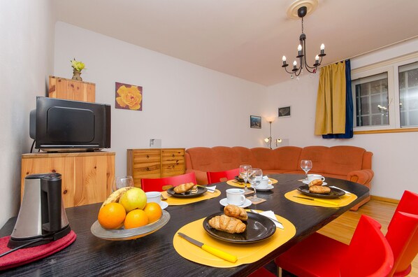 A dining area with a table set for breakfast, including croissants and oranges, and a television on a wooden cabinet. - Apartment Z4 (Split)