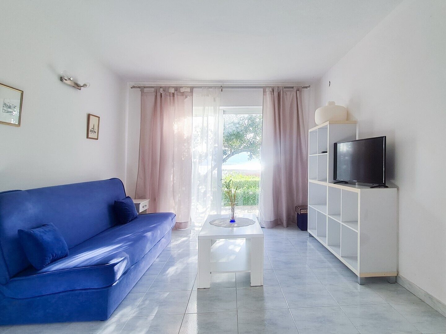 Un salon moderne avec un canapé bleu, une table basse blanche, un téléviseur posé sur un meuble blanc, et une vue sur la verdure grâce à de grandes fenêtres.