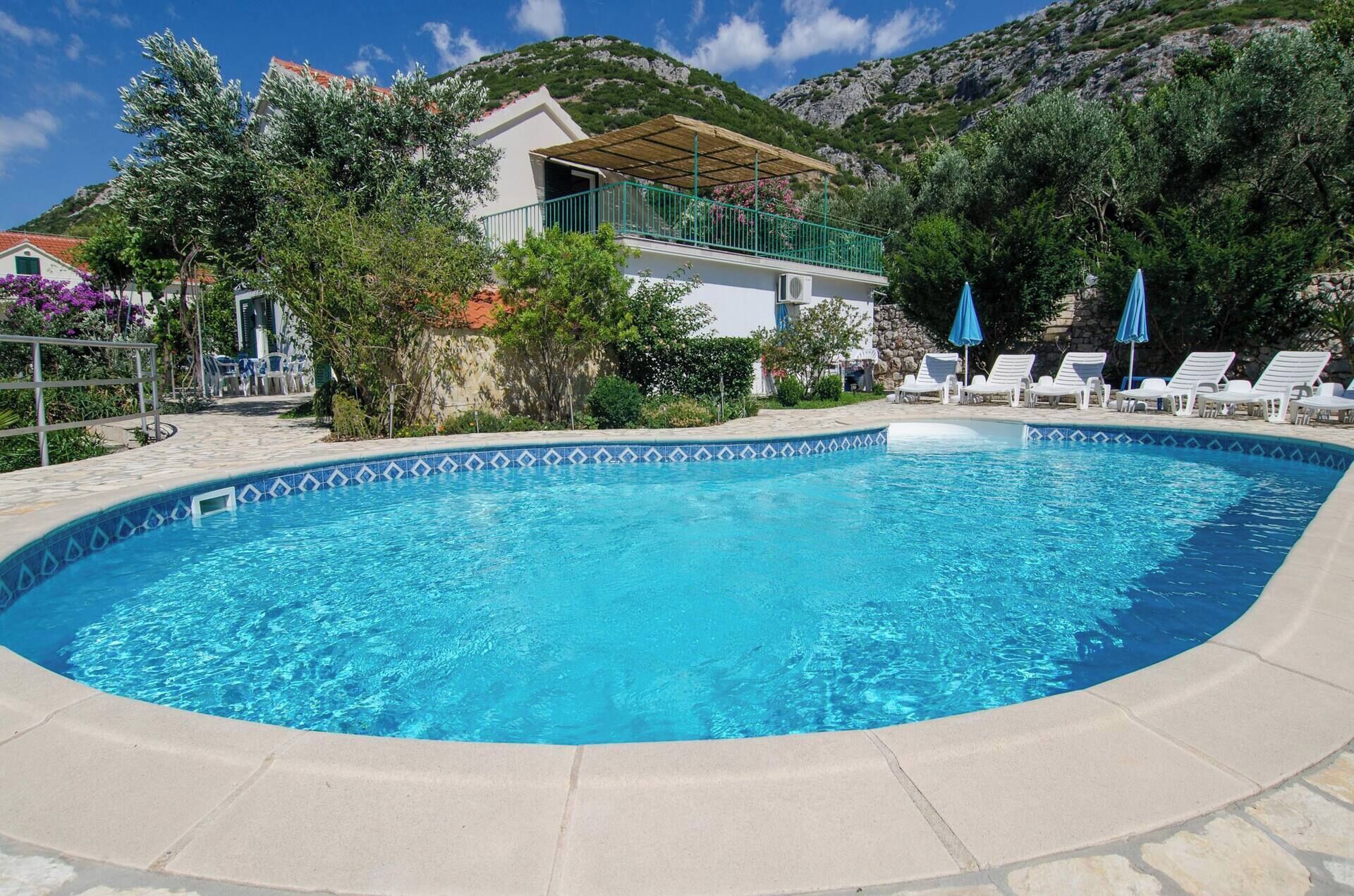 Ein Schwimmbecken mit gefliestem Rand, umgeben von Liegestühlen und Sonnenschirmen, vor einer Hügelkulisse und einem Gebäude mit Balkon.