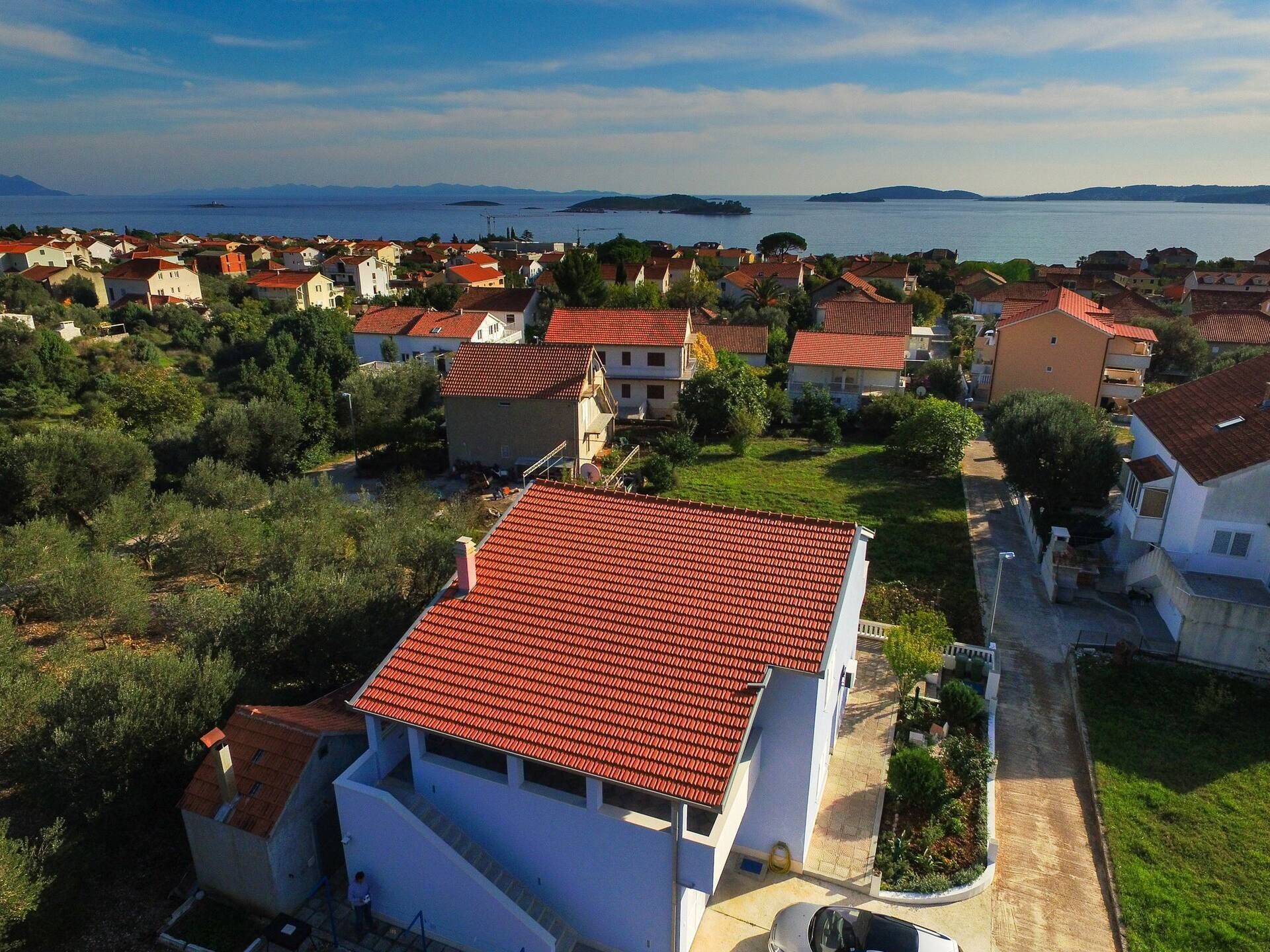 A coastal residential area with houses featuring red-tiled roofs, surrounded by greenery and a view of the sea.