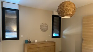 A bedroom with a bed, a wooden dresser, two windows with blinds, a woven pendant light, and a round wall decoration. - Seaside gîte 2 to 4 pers. (Vidauban)
