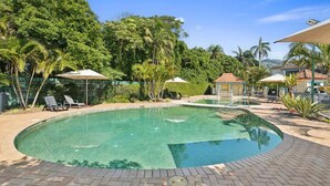 A clear swimming pool surrounded by lounge chairs and umbrellas, with lush greenery and a clear sky.