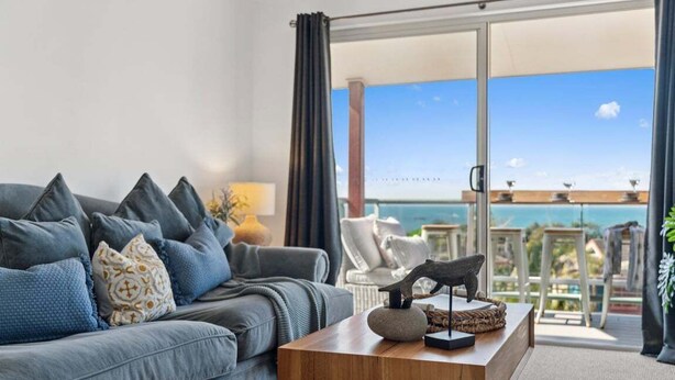 A modern living room with a grey sofa, a wooden coffee table, and a view of the ocean through a glass door.