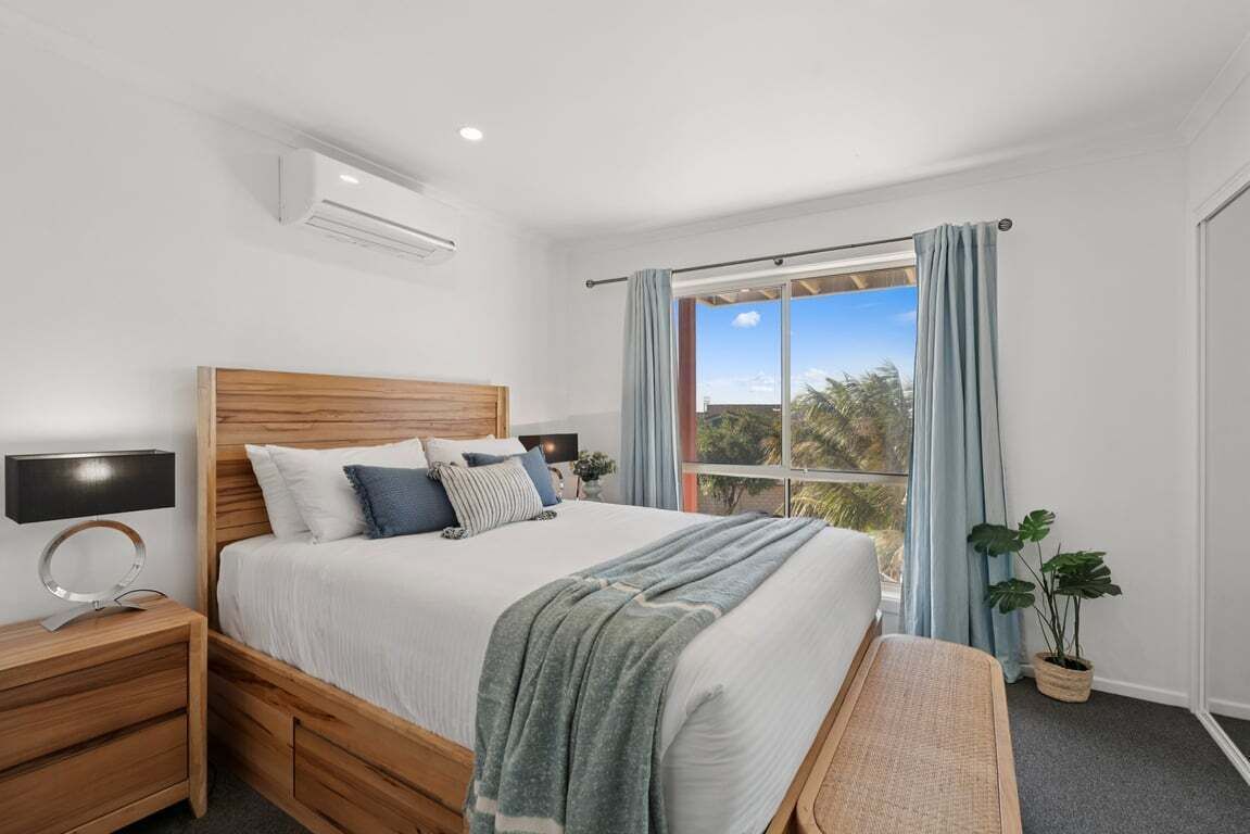 A bedroom with a wooden bed, a nightstand, a bedside lamp, a window with curtains, and a view of trees.