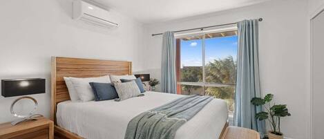 A bedroom with a wooden bed, a nightstand, a bedside lamp, a window with curtains, and a view of trees.