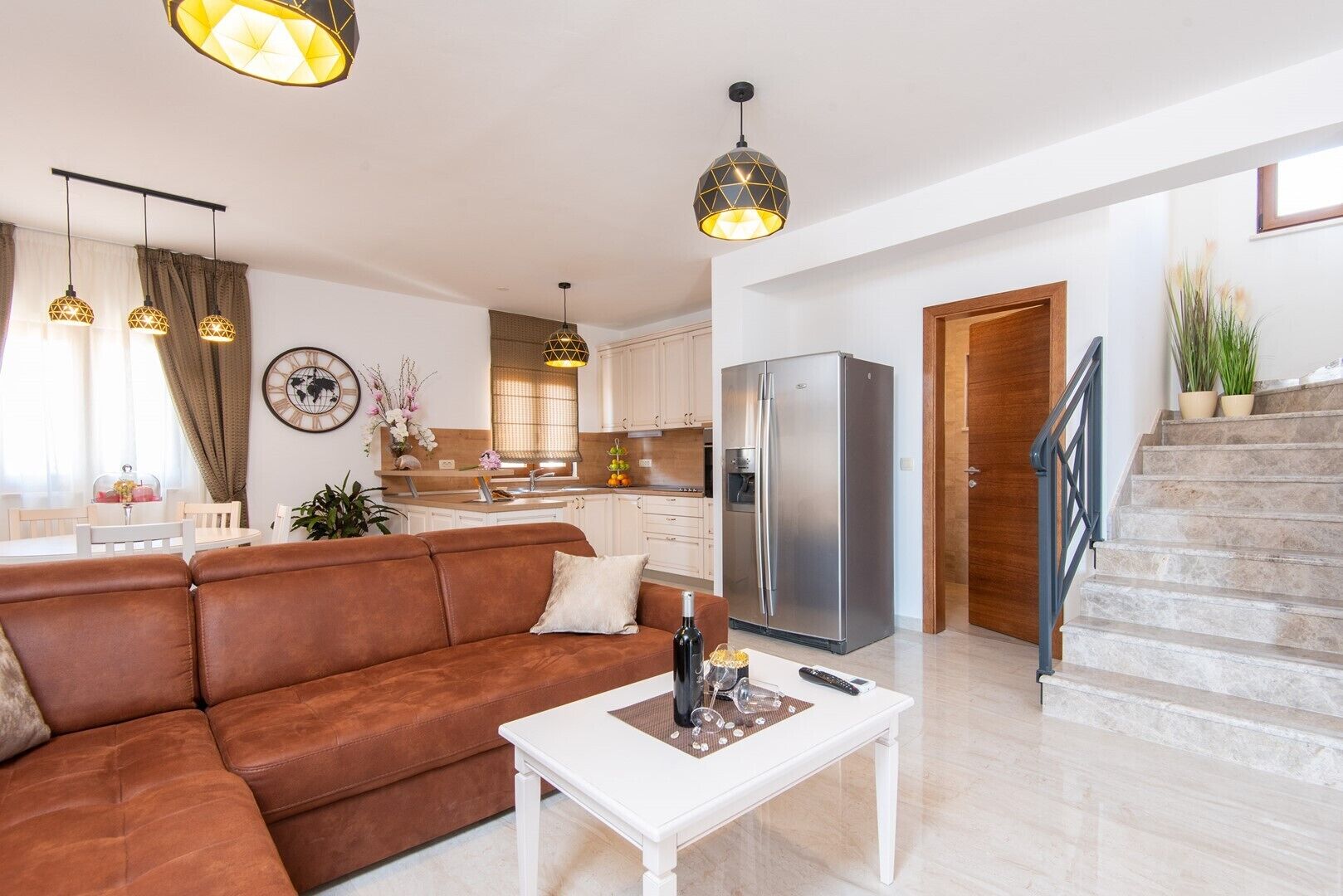 Una sala moderna con un sofá marrón, una mesa de centro blanca y lámparas colgantes. Se ve una cocina con armarios blancos y un refrigerador de acero inoxidable al fondo.