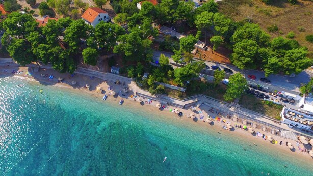 An aerial view of a beach with clear turquoise waters, a promenade lined with umbrellas, and buildings with red roofs.