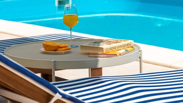 Una mesa junto a la piscina con un vaso de zumo de naranja, un plato de rodajas de naranja y un libro titulado 'The Daily Home Management'.