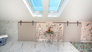 Un baño moderno con tragaluces, piso de mármol y un jarrón decorativo sobre una mesa de vidrio transparente.