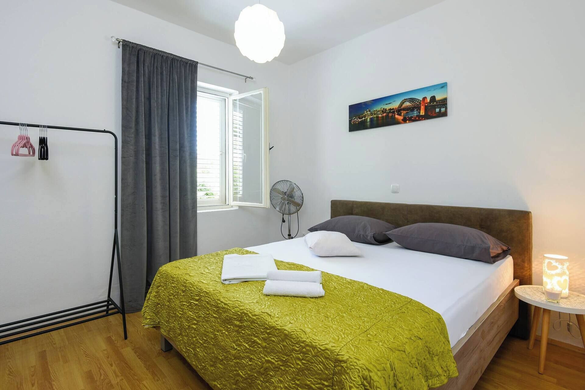 Une chambre d’hôtel avec un lit, une fenêtre avec des rideaux, un ventilateur, un tableau et une table de chevet avec une lampe.