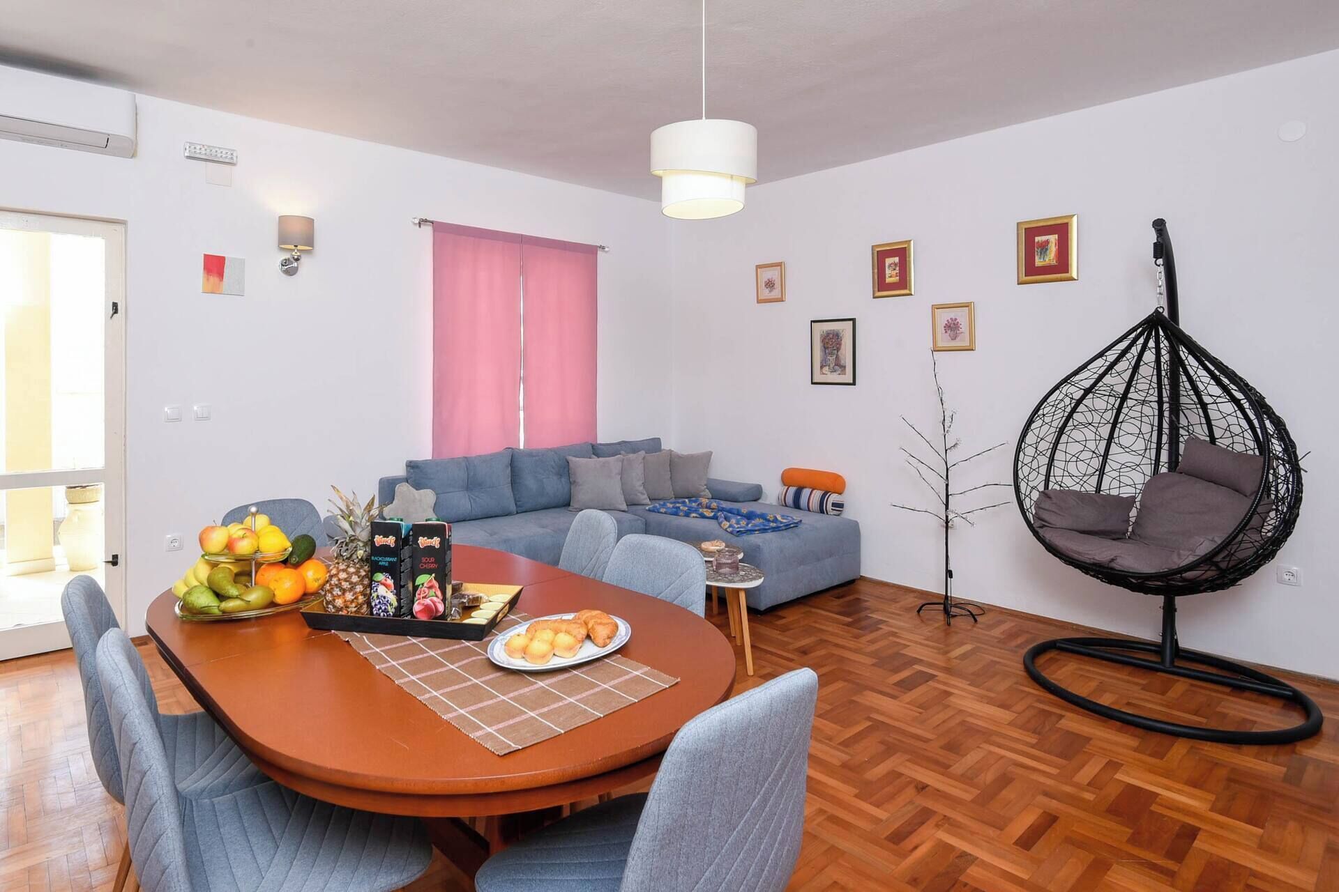 Una sala de estar moderna con una mesa de comedor, sofá gris, una silla colgante y un ventanal con cortinas rosadas.