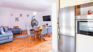 A modern living room with a sofa, dining table, chairs, and a hanging chair. A large refrigerator is visible in the kitchen area.