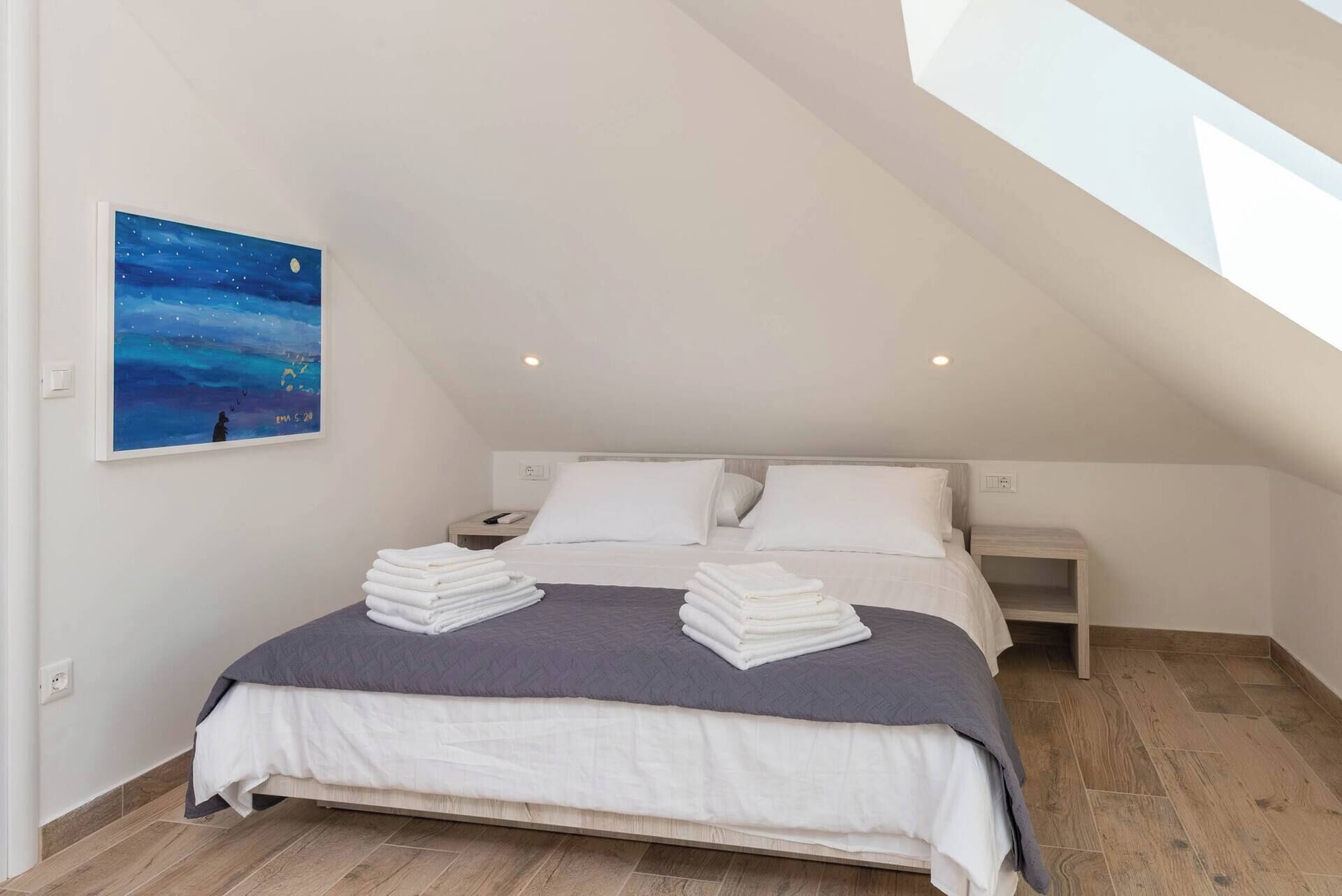 Een slaapkamer met schuine wand, een bed met witte beddengoed, twee kussens, een nachtkastje en een schilderij van een nachthemel.