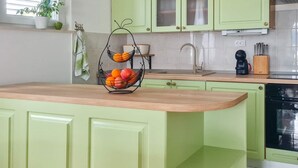 Una cocina con armarios verdes, encimera de madera, frutero y ventana con persianas.