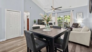 A modern dining area with a wooden table, black chairs, and a vase of flowers. There is a ceiling fan, a white couch, and a lamp in the background.