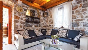 A cozy living room with a stone wall, a wooden ceiling, a black and white sofa, a glass coffee table, and a window with curtains. - The Hidden Treasure Village, 7-Bedroom Luxury Holiday Resort with children playground, private Pool and Private Parking, SEA VIEW (Orebic)