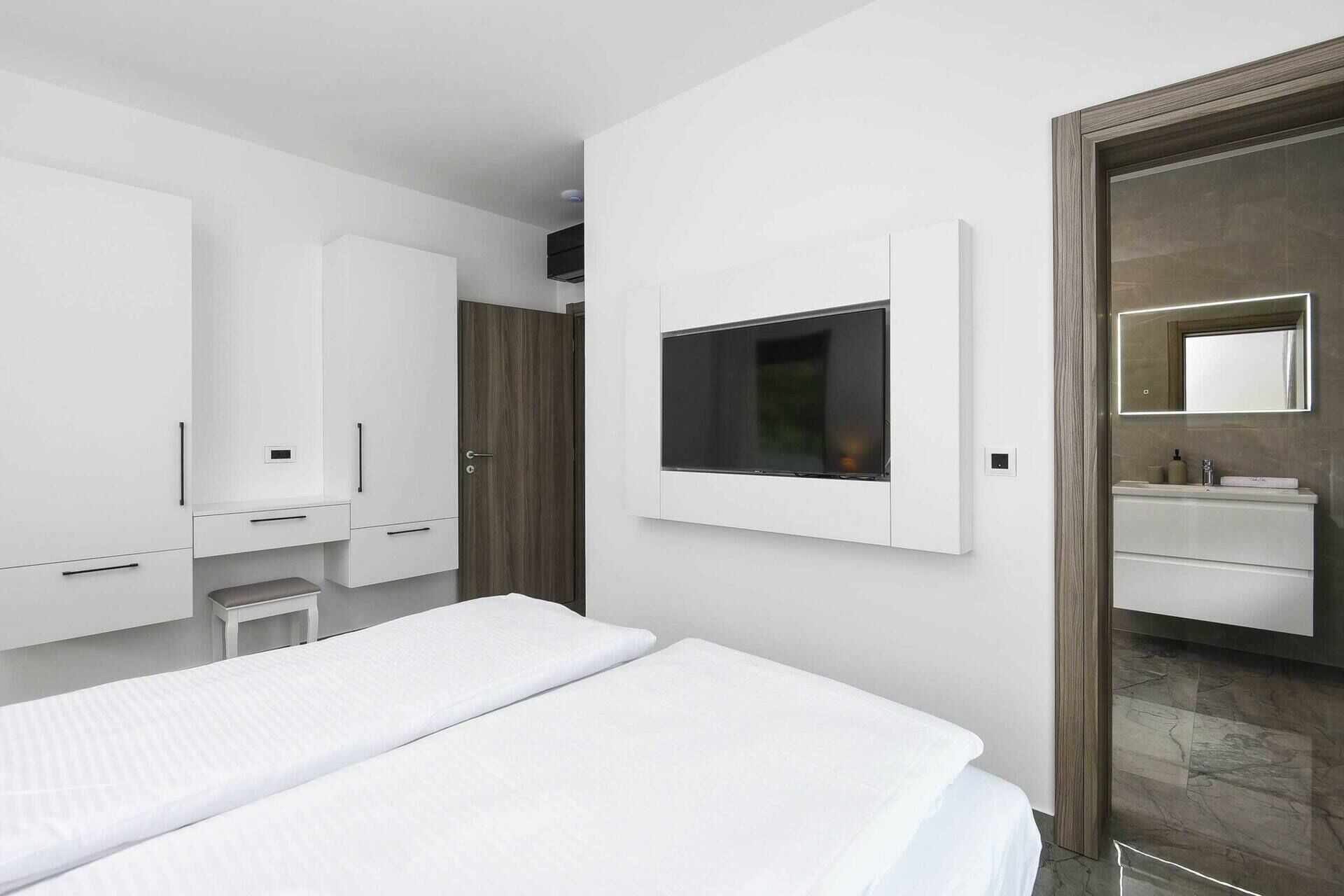 A modern bedroom with a bed, a TV mounted on the wall, a desk, and a bathroom with a mirror.