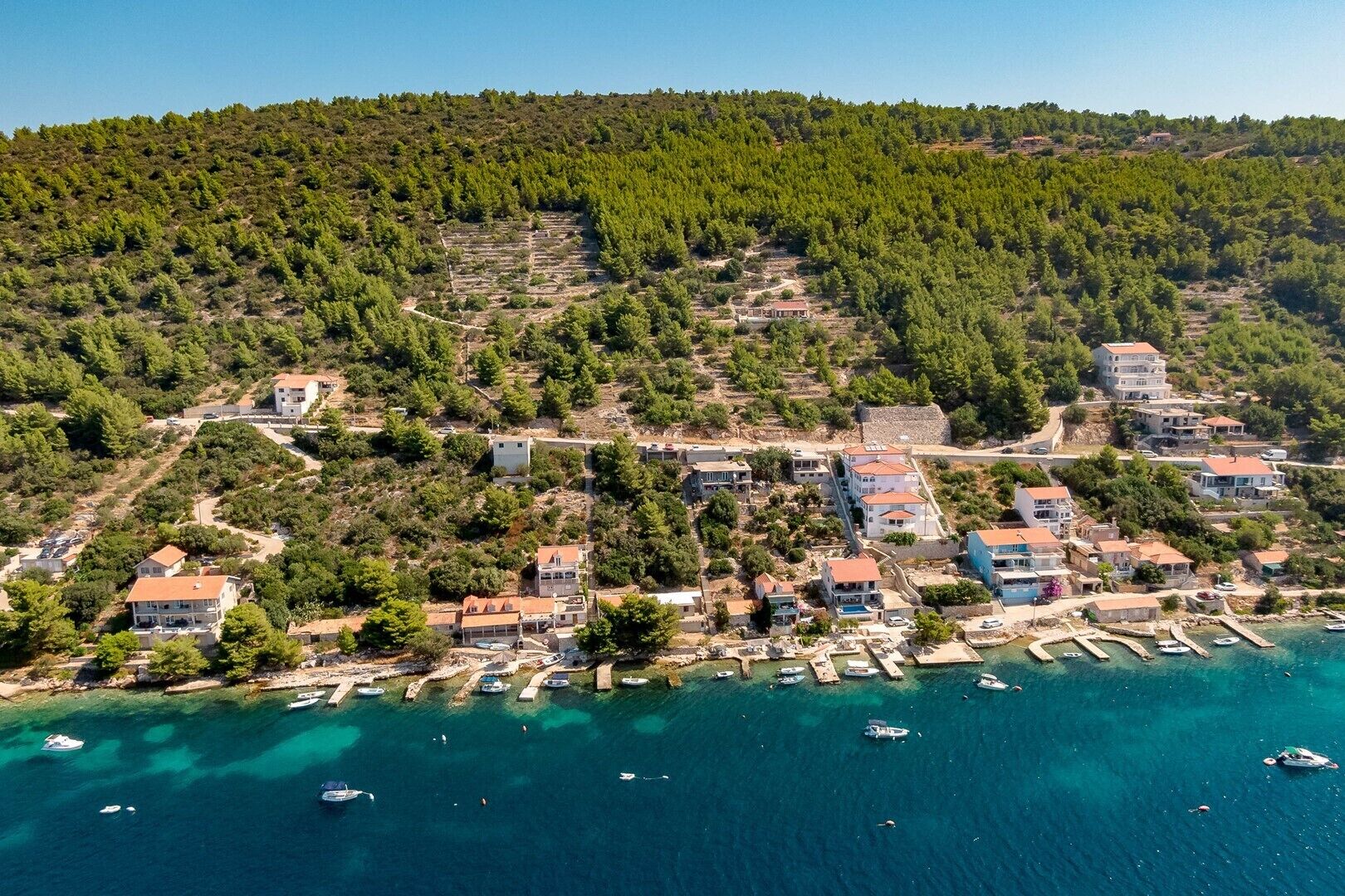 An aerial view of a coastal village with houses, a marina, and dense forested hills.