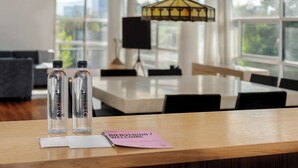 A modern dining area with a wooden table, two clear glass bottles labeled "POOMIK," a black lamp, and a "WELCOME" sign on a pink paper.