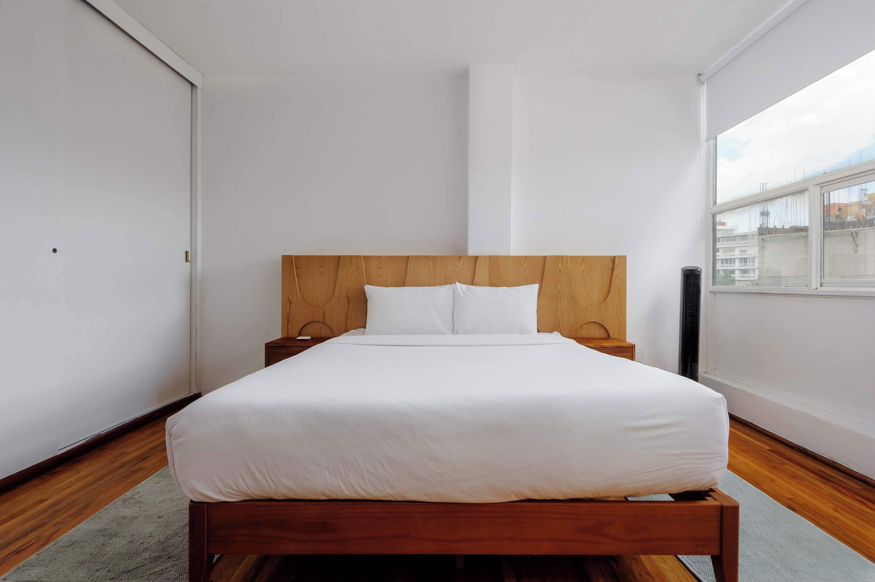 Een moderne slaapkamer met een houten bed, witte beddengoed, een nachtkastje en een raam met uitzicht over de stad.