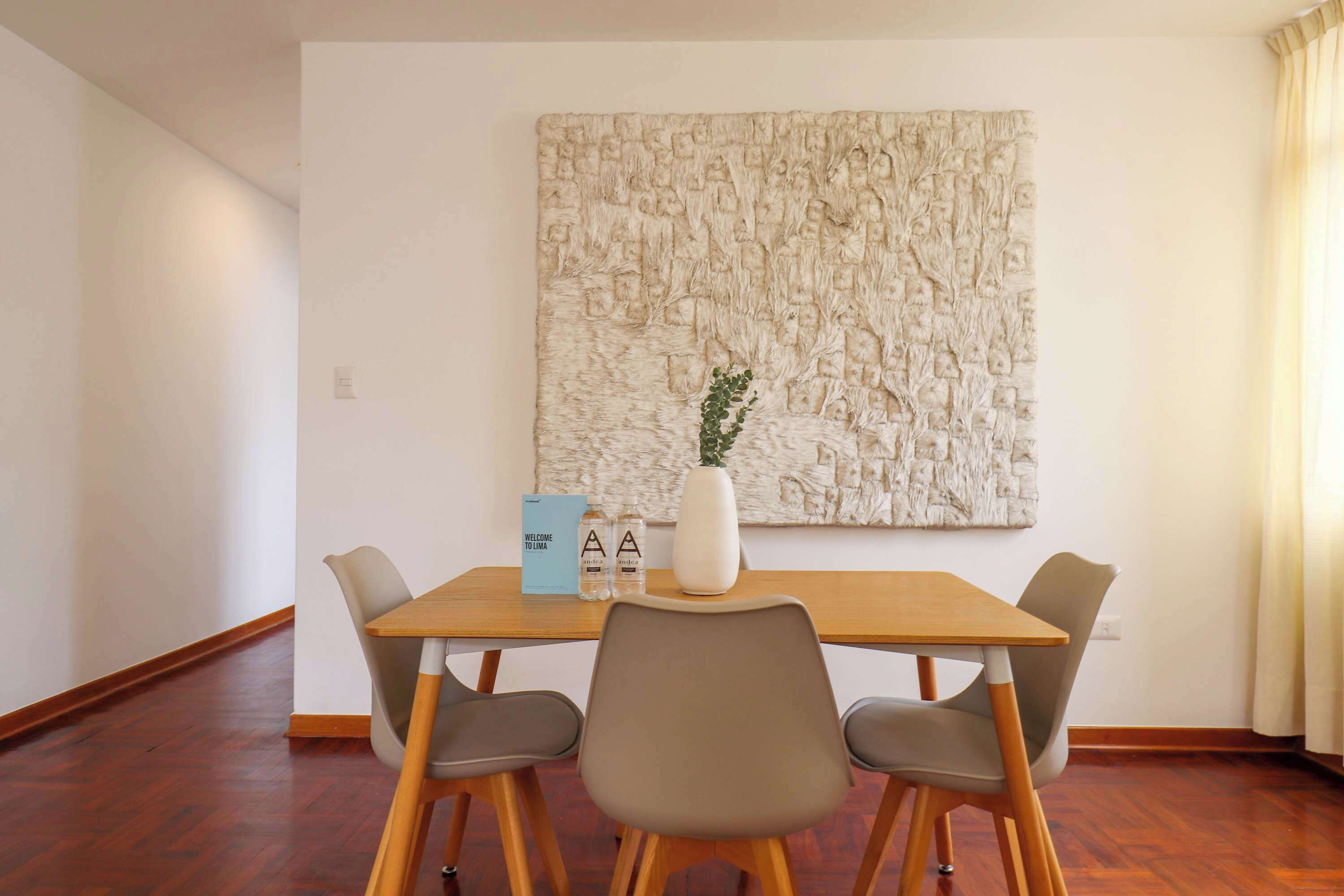 A dining area with a wooden table, four chairs, a vase with a plant, and a welcome sign.