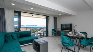 A modern living room with a green sofa, dining table, and chairs, a flat-screen TV, and a balcony with a view of the sea.